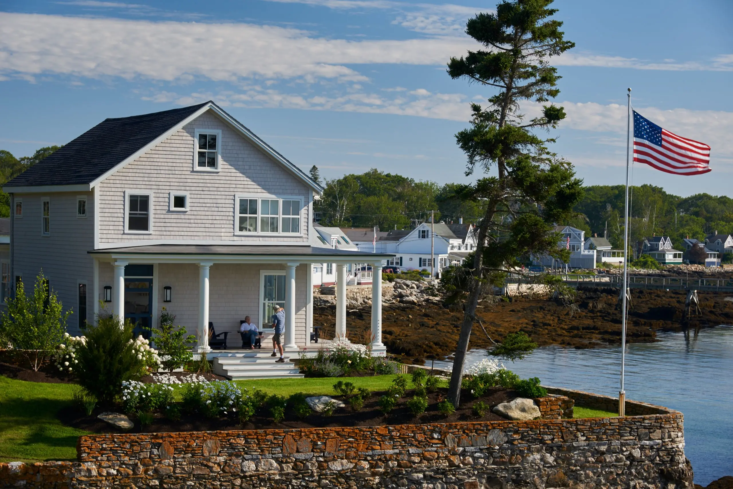 Welcoming view of coastal Maine home with flag, stone wall, classic pillars, a wrap porch, and beautiful floral accent along the ocean.